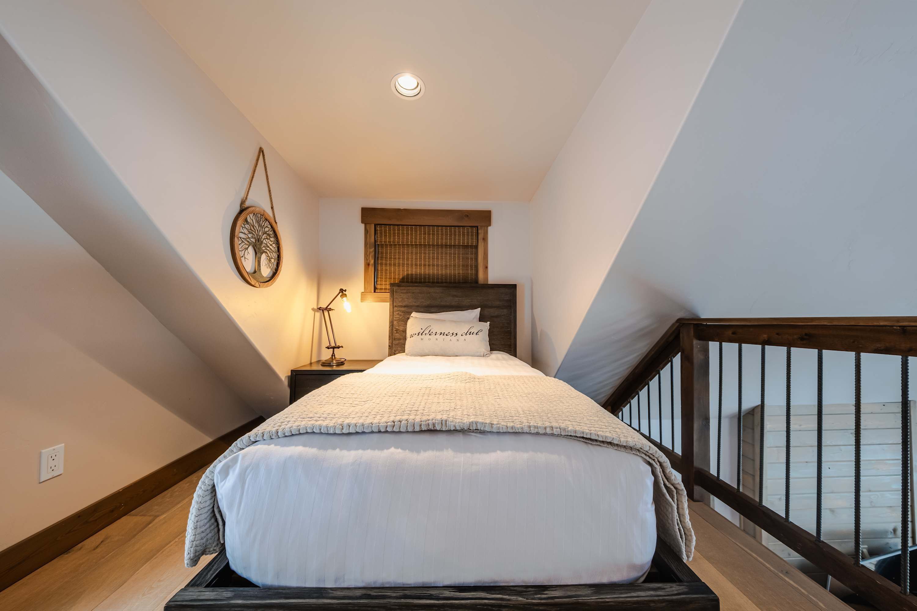 A bedroom with a bed, a nightstand, a wall-mounted lamp, a window with a wooden blind, and a wooden railing.