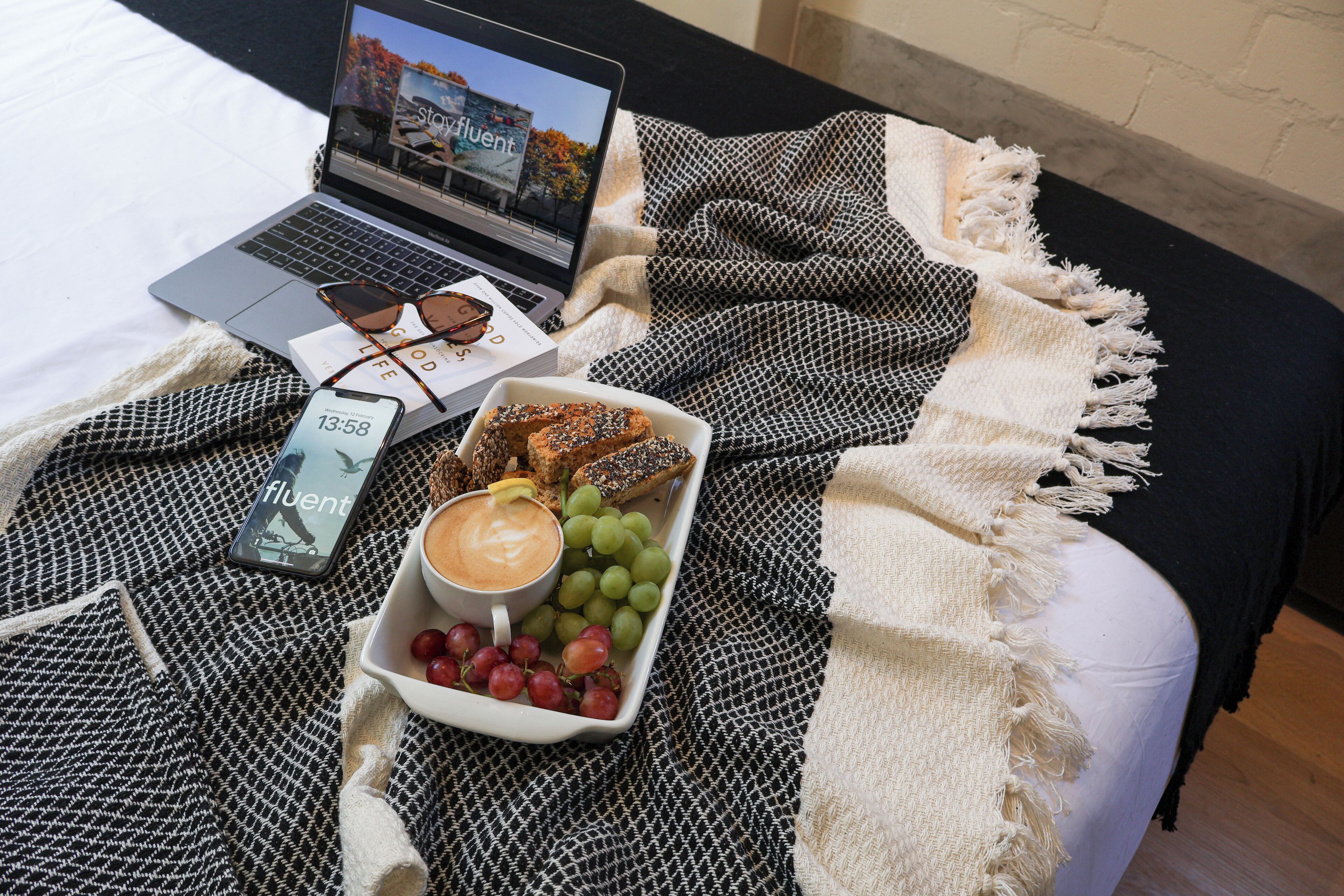 A laptop on a bed with a blanket, a tray with snacks, and a smartphone displaying the time.