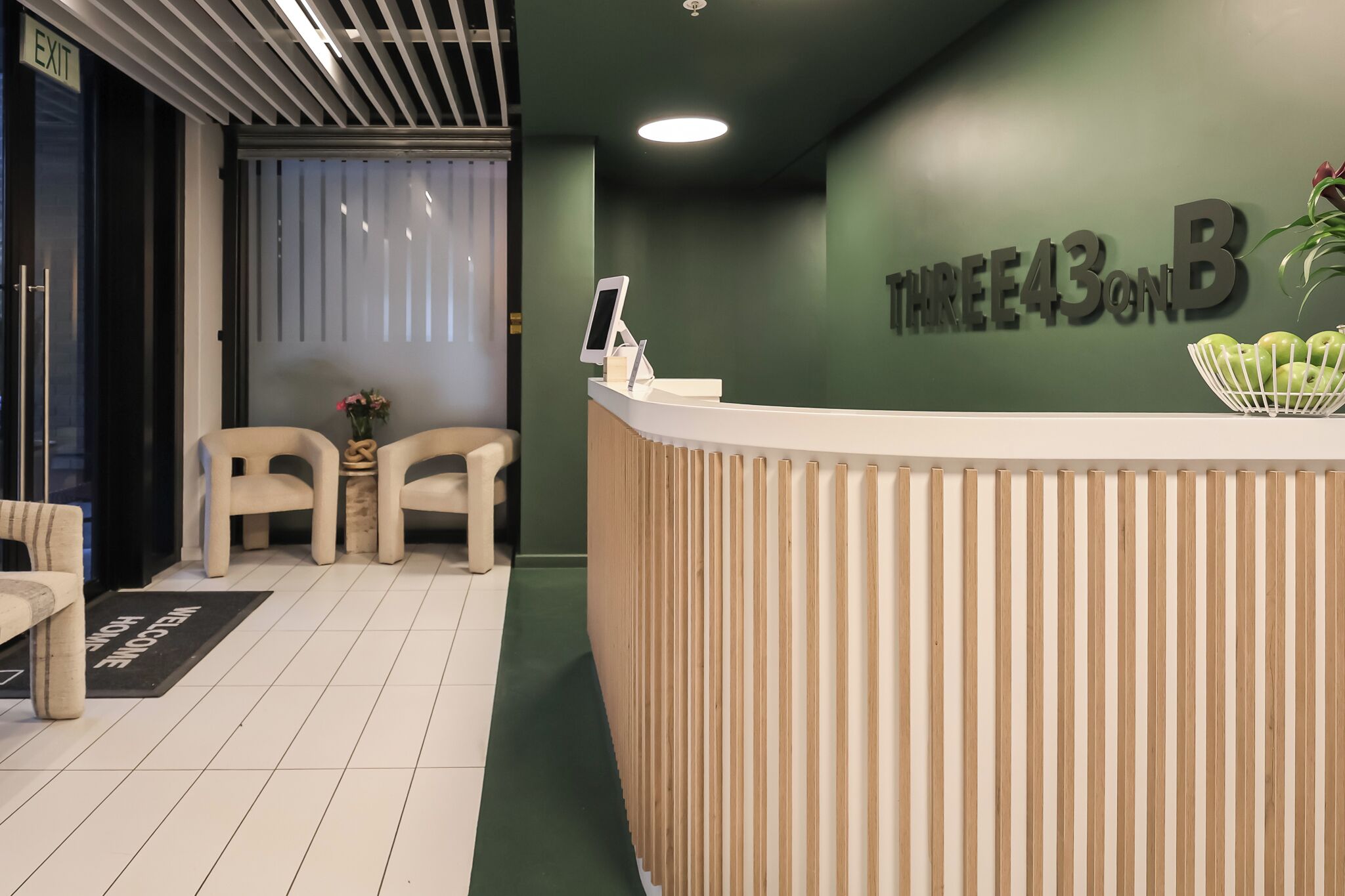 A reception desk with a sign reading 'THREE43ONB' and a plant on top. There are two chairs and a small table with a vase of flowers.