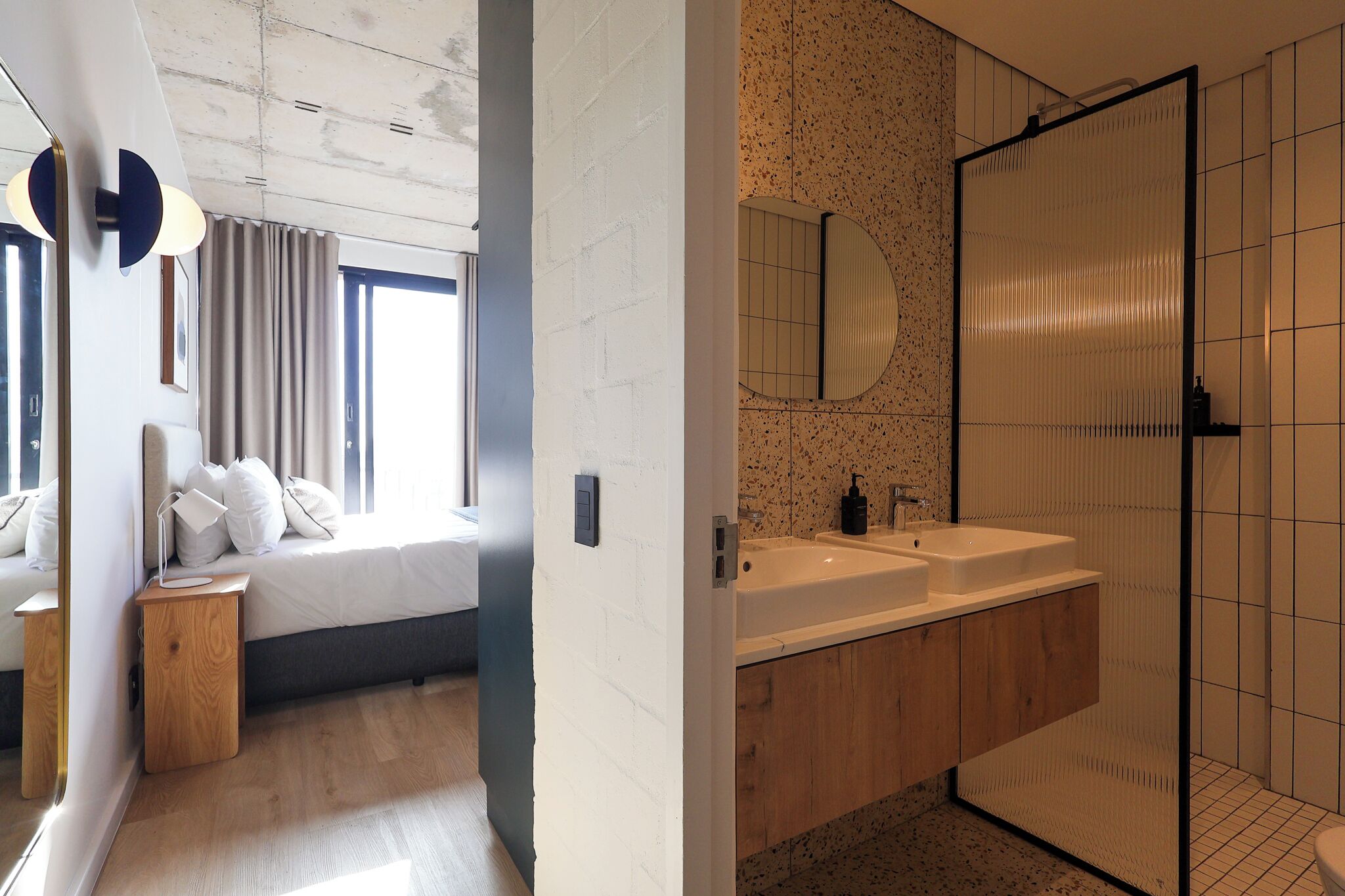 Une chambre moderne avec un grand lit, une table de chevet et un miroir. À côté se trouve une salle de bain avec un double lavabo, un miroir et un espace douche.