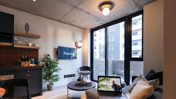 Un appartement moderne comprenant une cuisine, un coin repas et un salon. Il est doté d’une grande fenêtre, d’un téléviseur à écran plat et d’un ordinateur portable posé sur le canapé.