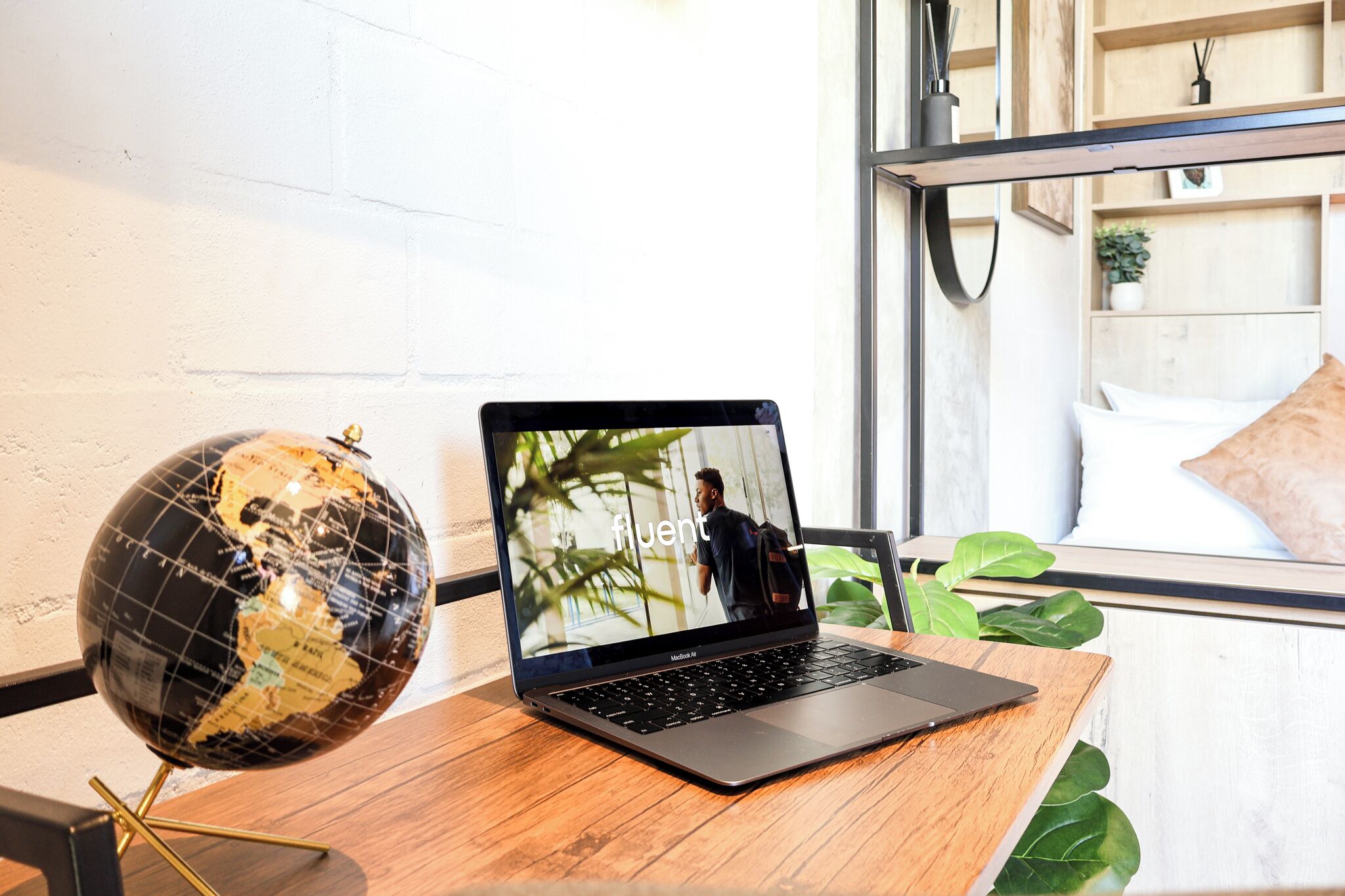 A laptop on a wooden table with a globe beside it, a bed with a white pillow, and a window with a view of greenery.