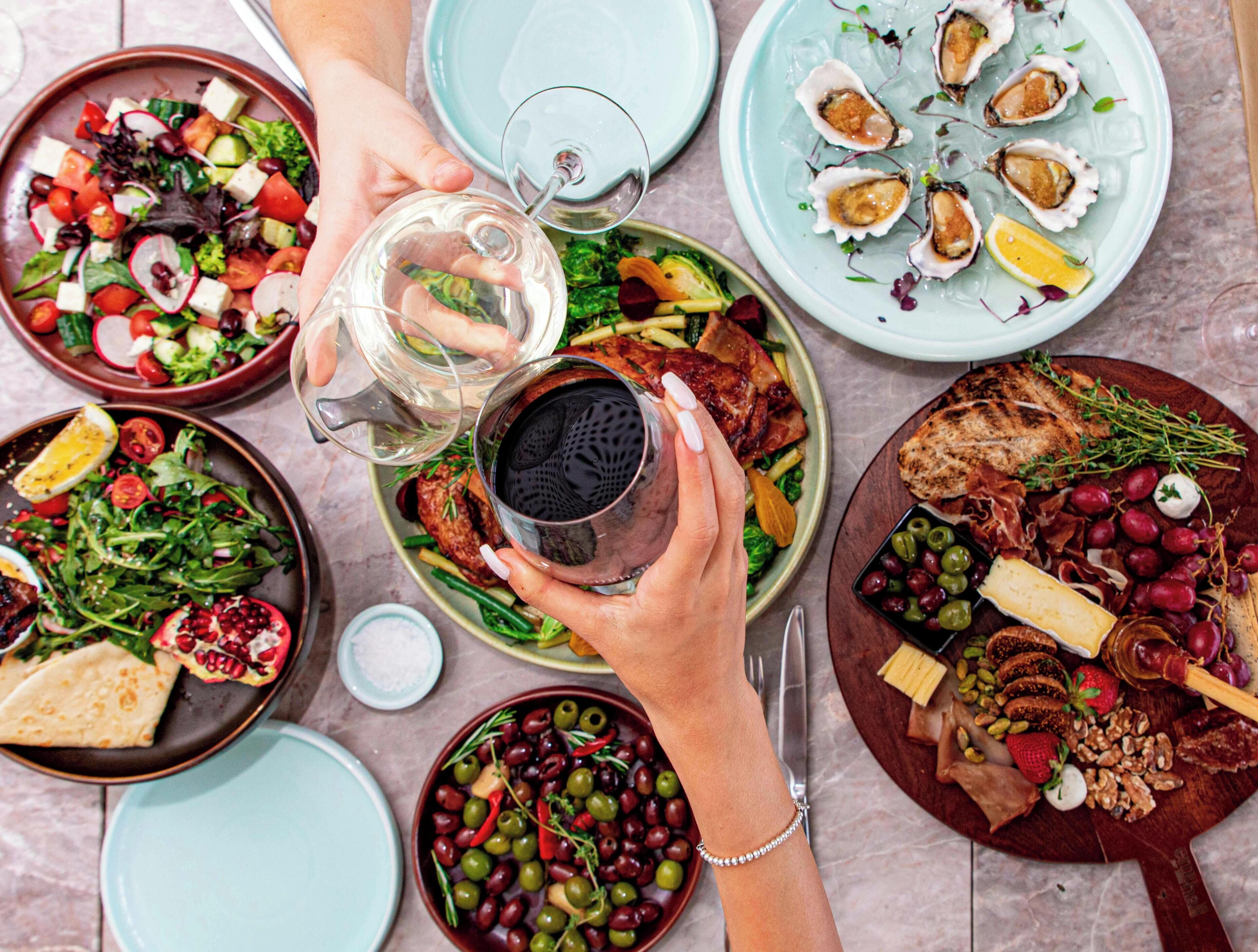 Diverse gerechten op een tafel, zoals salades, vleeswaren en kazen, terwijl iemand een drankje inschenkt.