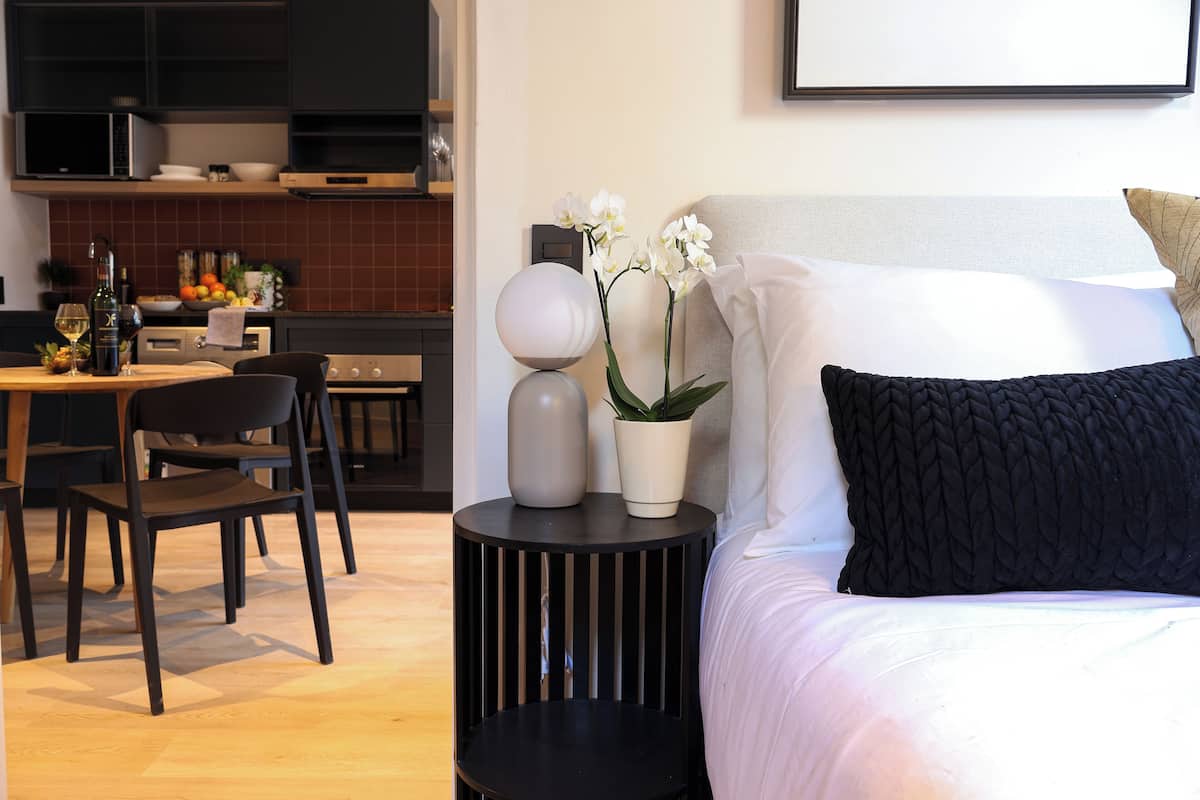 A modern bedroom with a bed, a bedside table with decorative items, a dining area with chairs and a table, and a kitchen in the background.