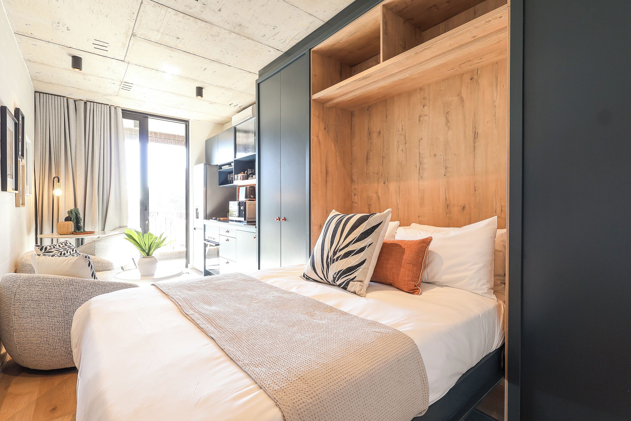A modern bedroom with a large bed, a wooden headboard, and a view into the kitchen area.
