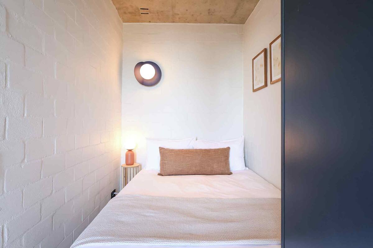 A narrow room with a bed, a round wall light, and framed pictures on the wall.