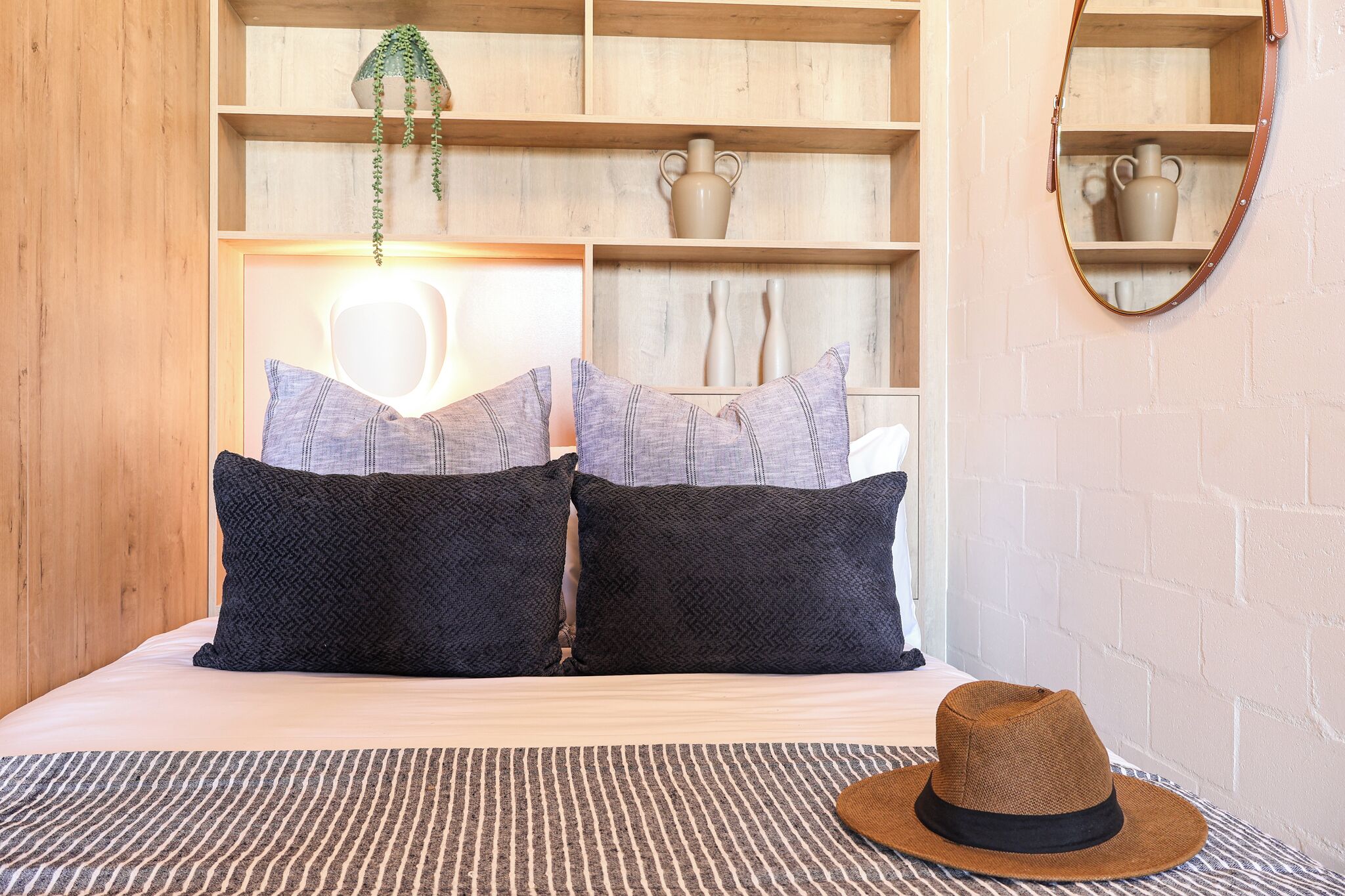 A neatly made bed with two dark pillows, a striped rug, and a straw hat on the side.