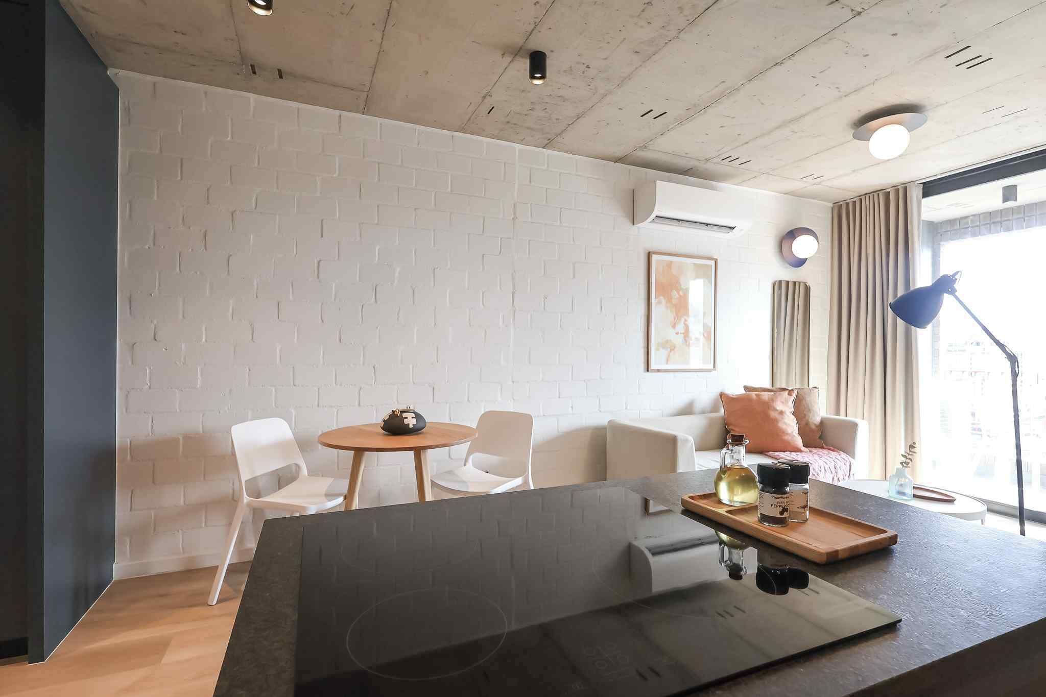 A modern room with a white brick wall, a round table with chairs, a sofa with cushions, a desk with a tray, and a window with curtains.