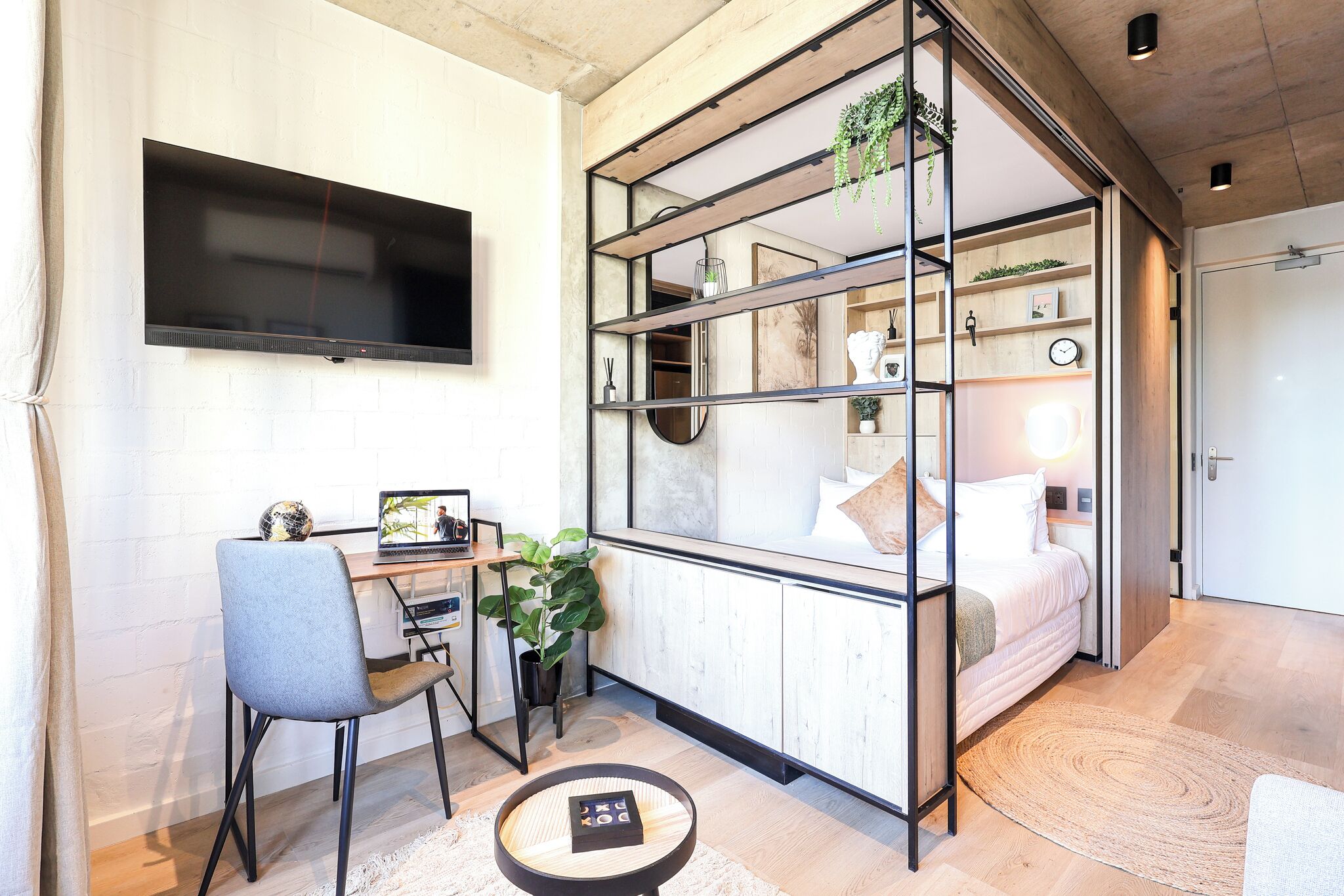 Habitación pequeña con cama, escritorio y silla, televisión en la pared y una planta.