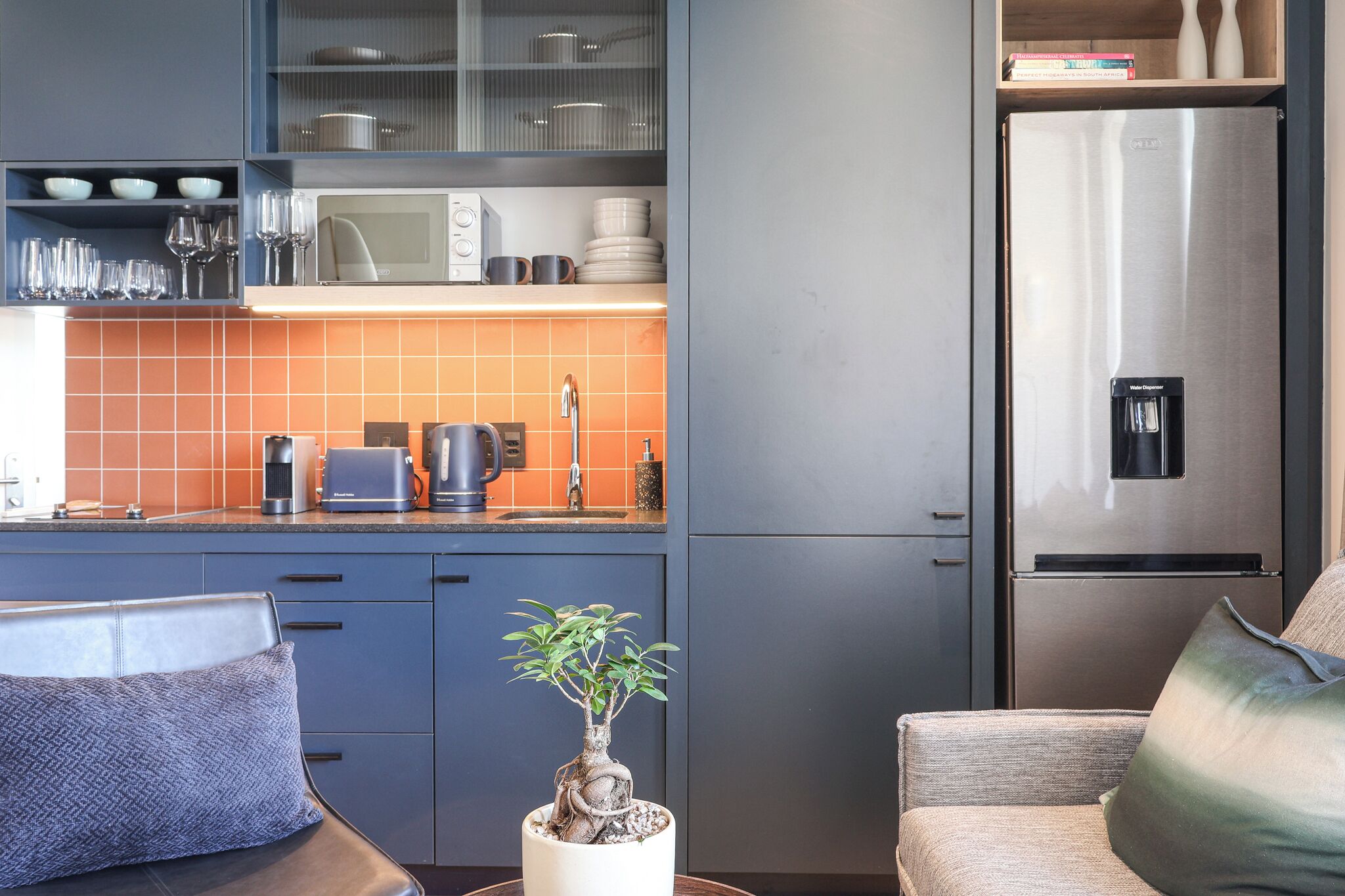 Una cocina moderna con microondas integrado, cafetera y hervidor. Hay un refrigerador, una planta en maceta y una zona de estar con sofá y cojines.