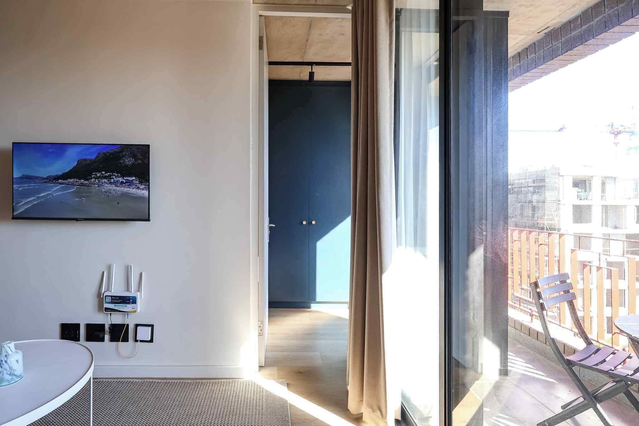 A room with a TV mounted on the wall, a small table, and a chair. There is a door leading to a balcony with a view of buildings.