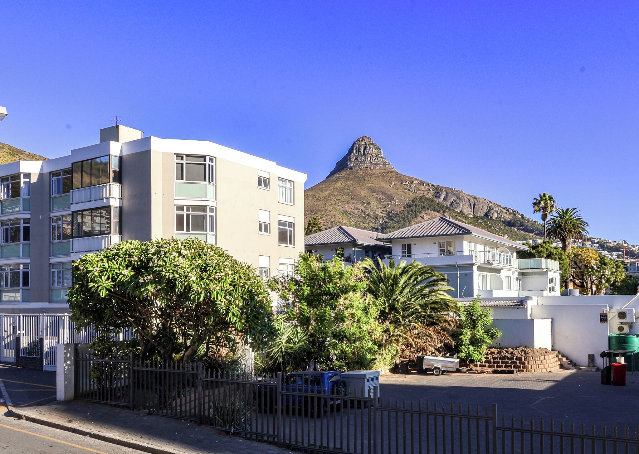 A modern building with multiple floors and balconies, situated in a residential area with lush greenery and a prominent mountain in the background.