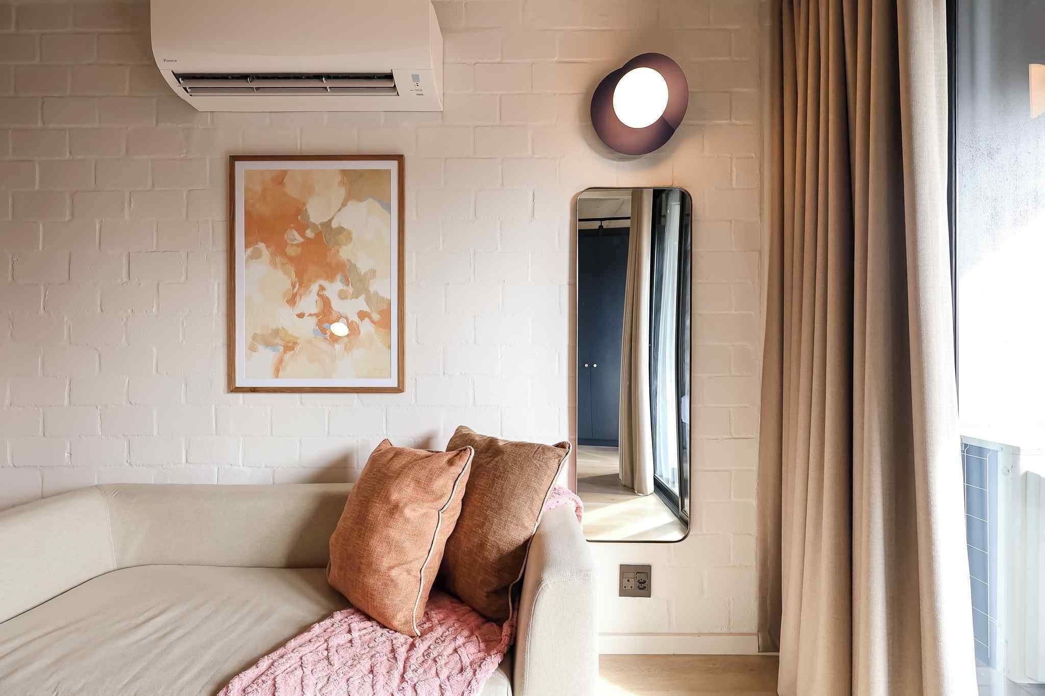 Ein Hotelzimmer mit weißer Ziegelwand, Klimaanlage, einem gerahmten abstrakten Gemälde, einem Spiegel, einem Fenster mit Vorhängen und einem beigefarbenen Sofa mit zwei Kissen.