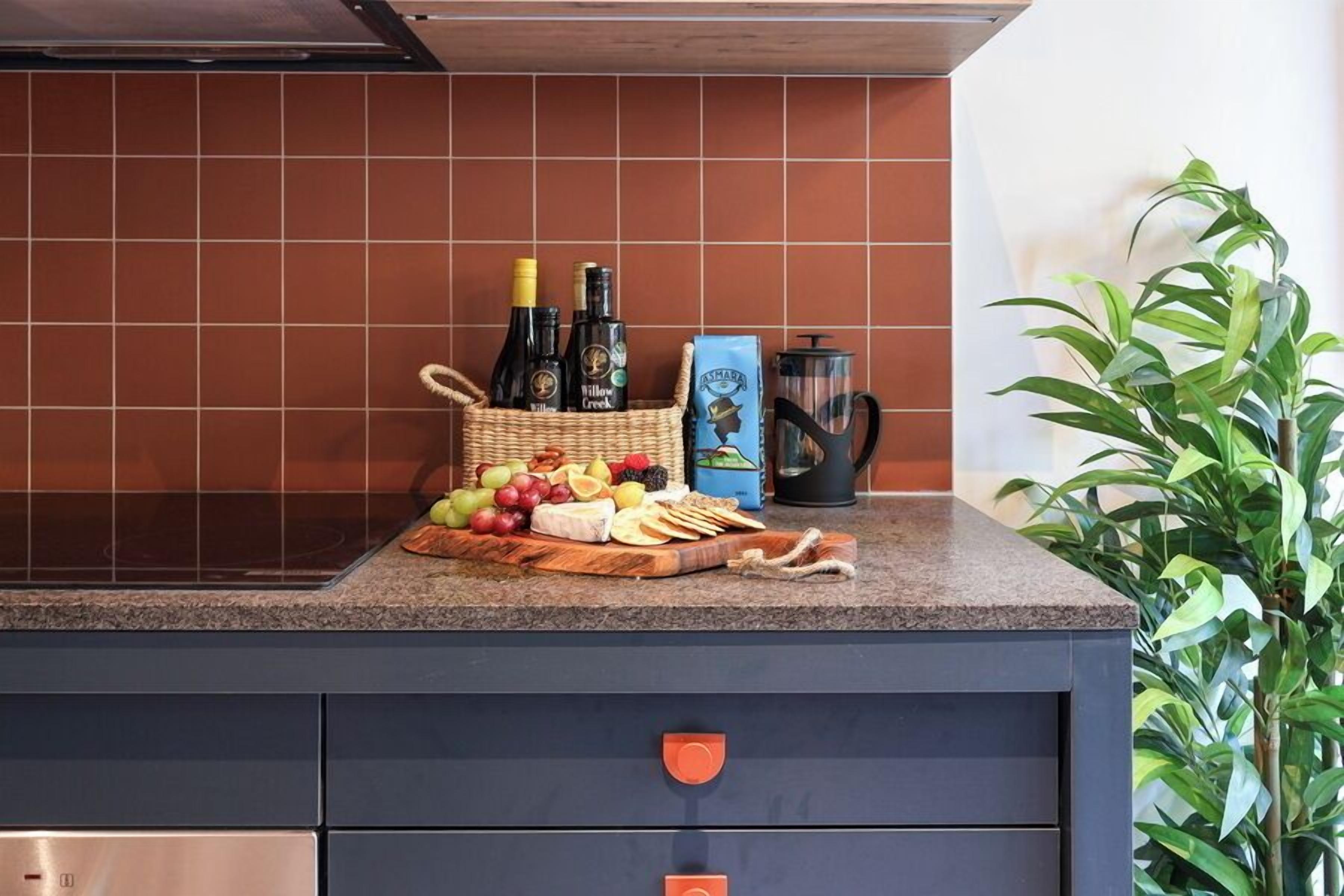 A kitchen counter with a fruit and cheese board, bottles, a coffee maker, and a basket.