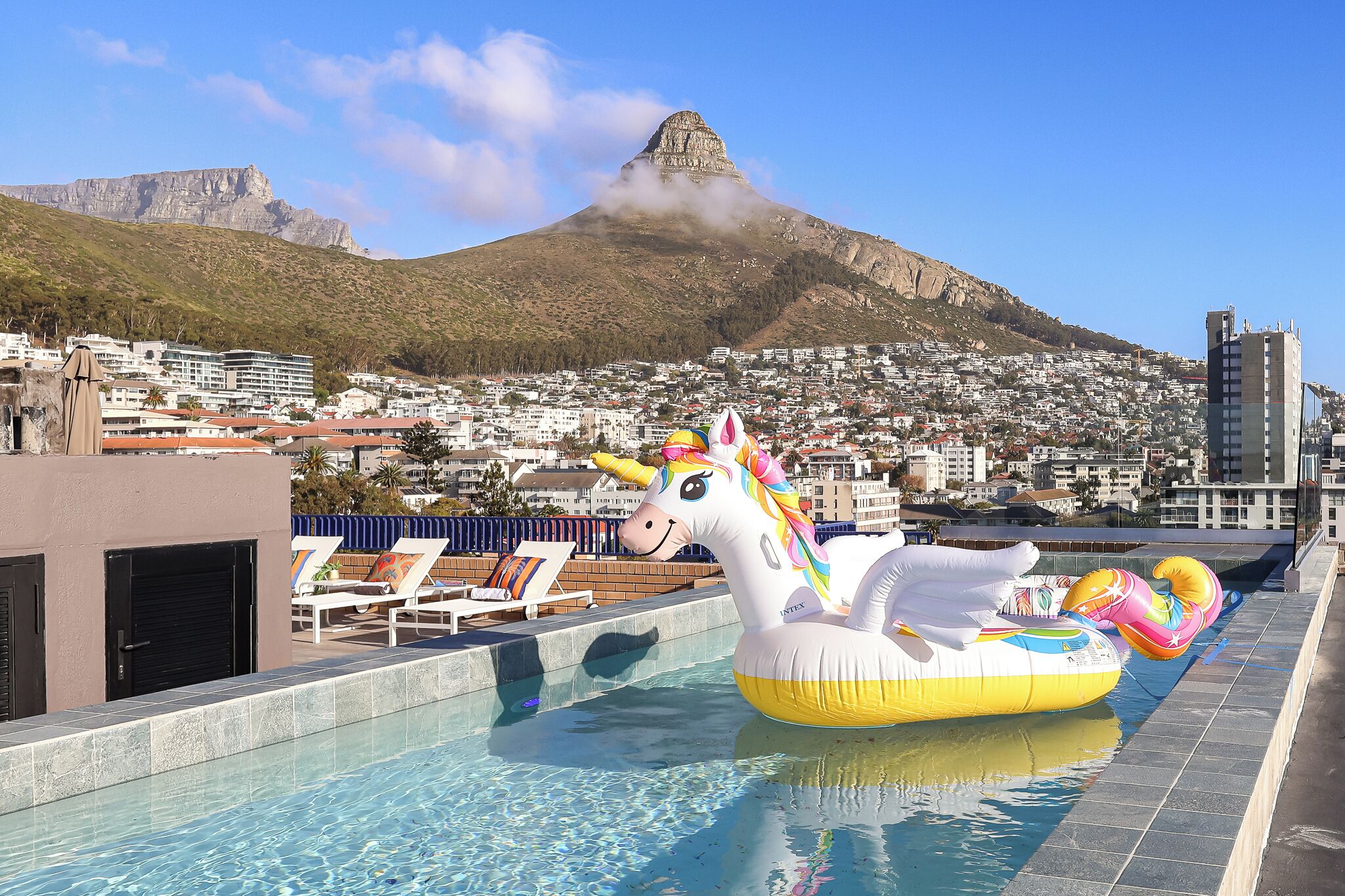 A swimming pool with inflatable unicorn floats, lounge chairs, and a view of a mountain and cityscape.
