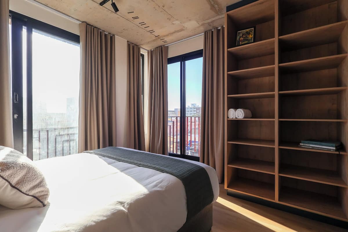 A hotel room with a large bed, a wooden bookshelf, and a view of the city through the windows.