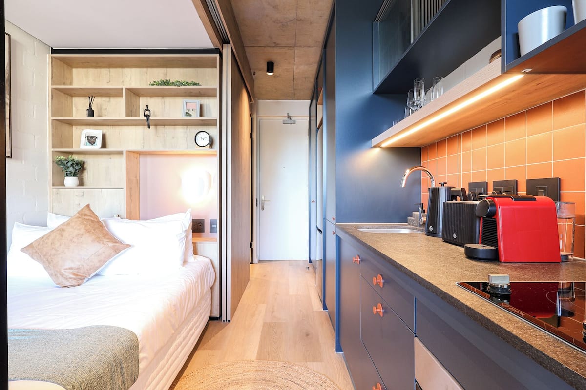 A modern hotel room with a bed, bedside table, and a built-in kitchen area with a sink, stove, and cabinets.