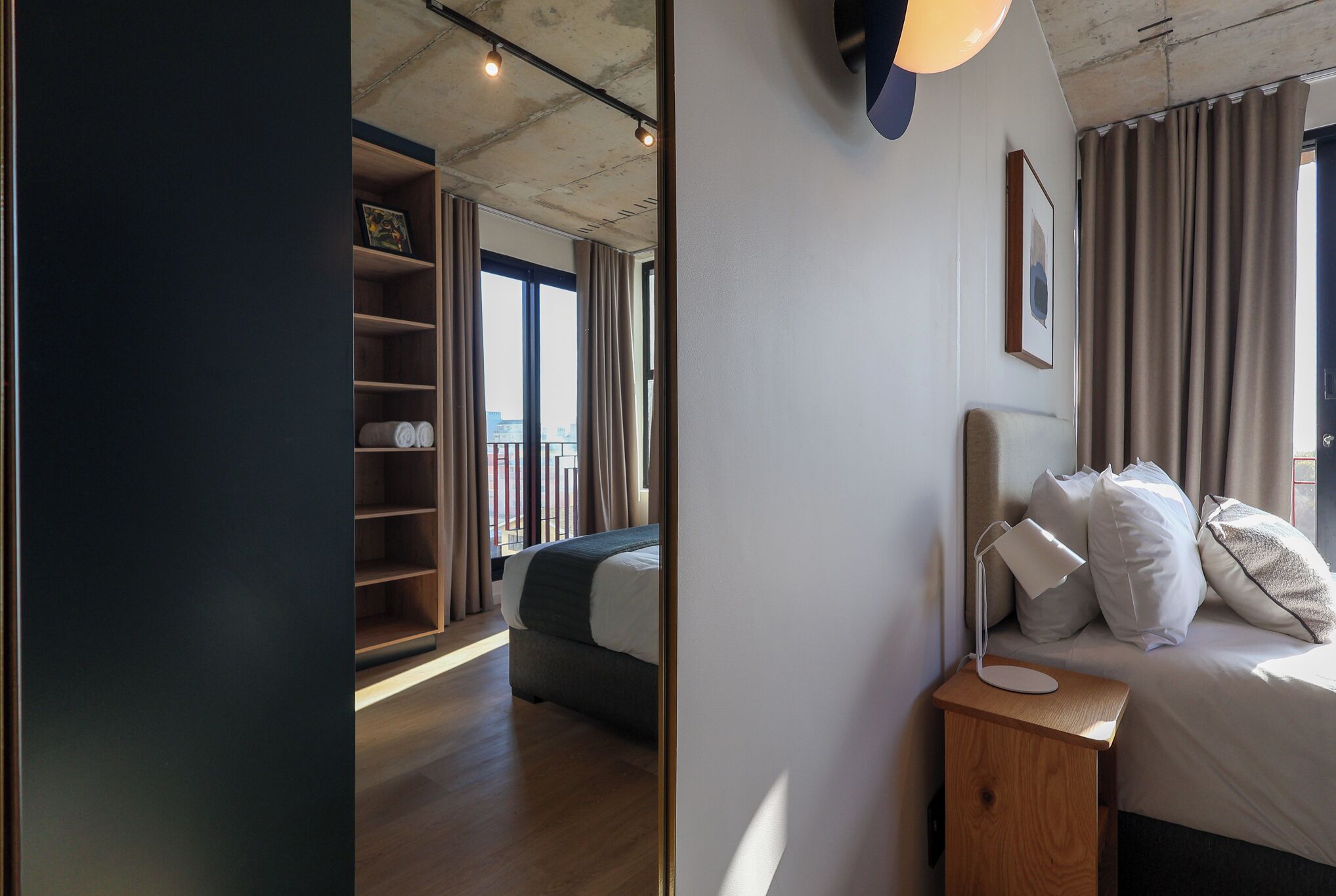 Une chambre d’hôtel moderne avec un plancher en bois, un lit avec une literie blanche, une table de chevet avec une lampe et une vue sur la ville grâce à de grandes fenêtres.