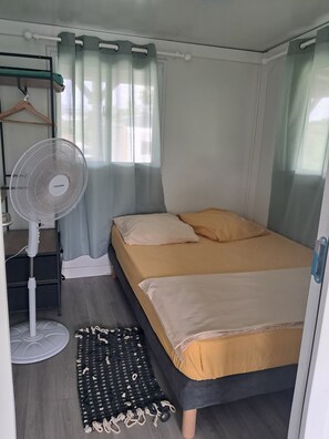 Ein kleines Schlafzimmer mit einem Bett, einem Standventilator, einem Teppich und einem Fenster mit Vorhängen.