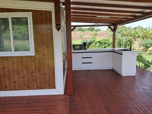 A covered outdoor kitchen area with a wooden deck, a white built-in kitchen counter, and a view of a lush garden.