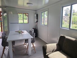 A modern kitchen with a white table, chairs, and a refrigerator. There are windows providing natural light and views of greenery outside.
