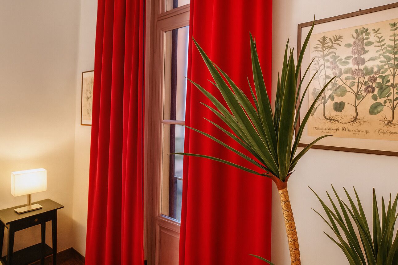A room with a window, red curtains, a potted plant, and a framed botanical illustration on the wall.