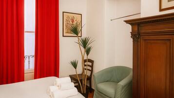 Quarto de hotel com cama, poltrona verde, aparador de madeira e uma planta.