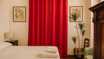 Quarto de hotel com cama, cortinas vermelhas, mesa de cabeceira com abajur, duas ilustrações botânicas emolduradas e uma planta em vaso.