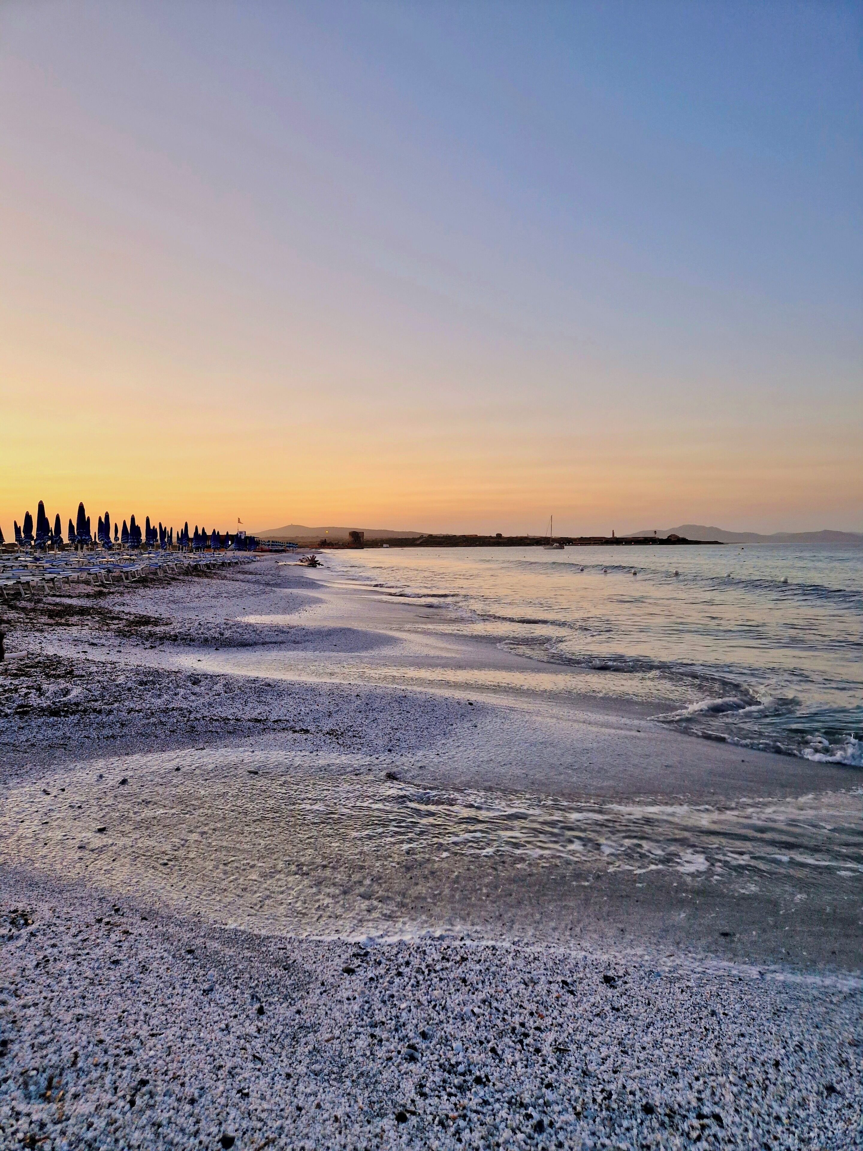 A beach with pebbles and waves, a distant shoreline with trees, and a clear sky.