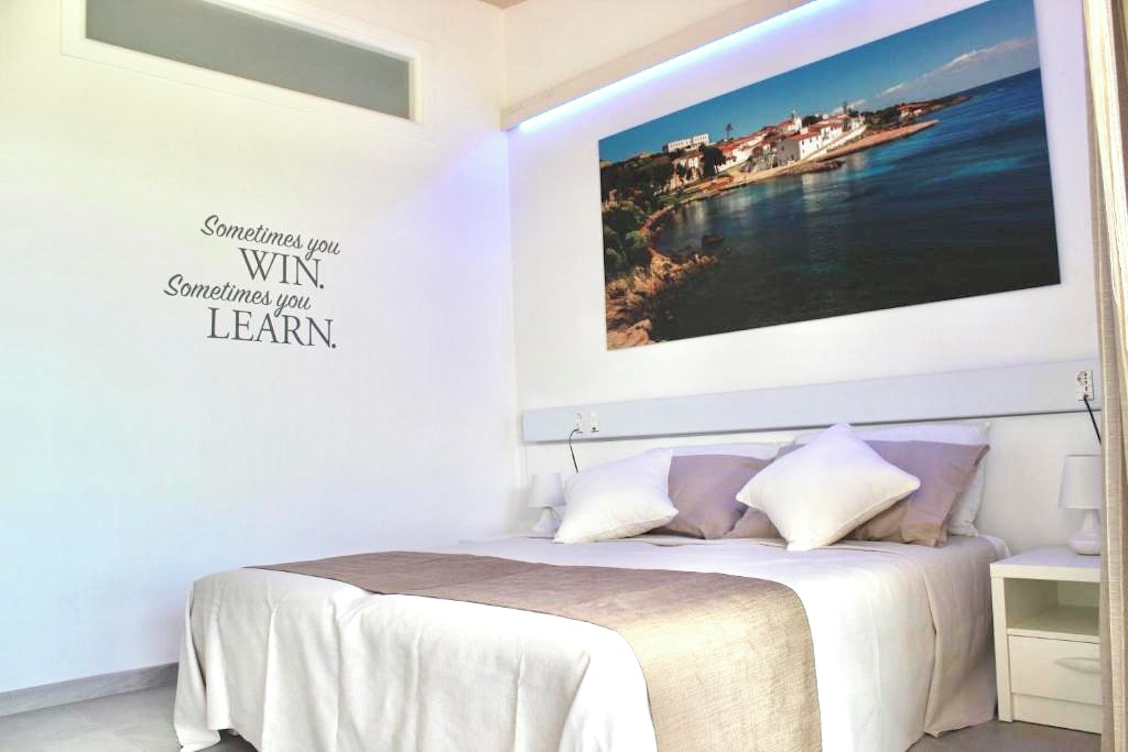Un dormitorio moderno con una cama grande, una mesita de noche y un televisor montado en la pared que muestra una escena costera. Se aprecia una frase motivacional en la pared.