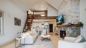 A spacious living room with a fireplace, a staircase leading to an upper level, and a kitchen in the background.