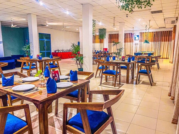 Un grand espace dédié à la salle à manger, avec plusieurs tables et chaises, dont certaines sont dotées de coussins bleus. On peut apercevoir, au fond, des plantes en pot et un écran de télévision.
