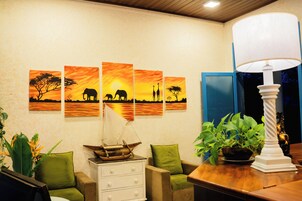 A room with a wall-mounted painting of a sunset scene, a wooden ship model, green cushioned chairs, a white dresser, and a table with a lamp.