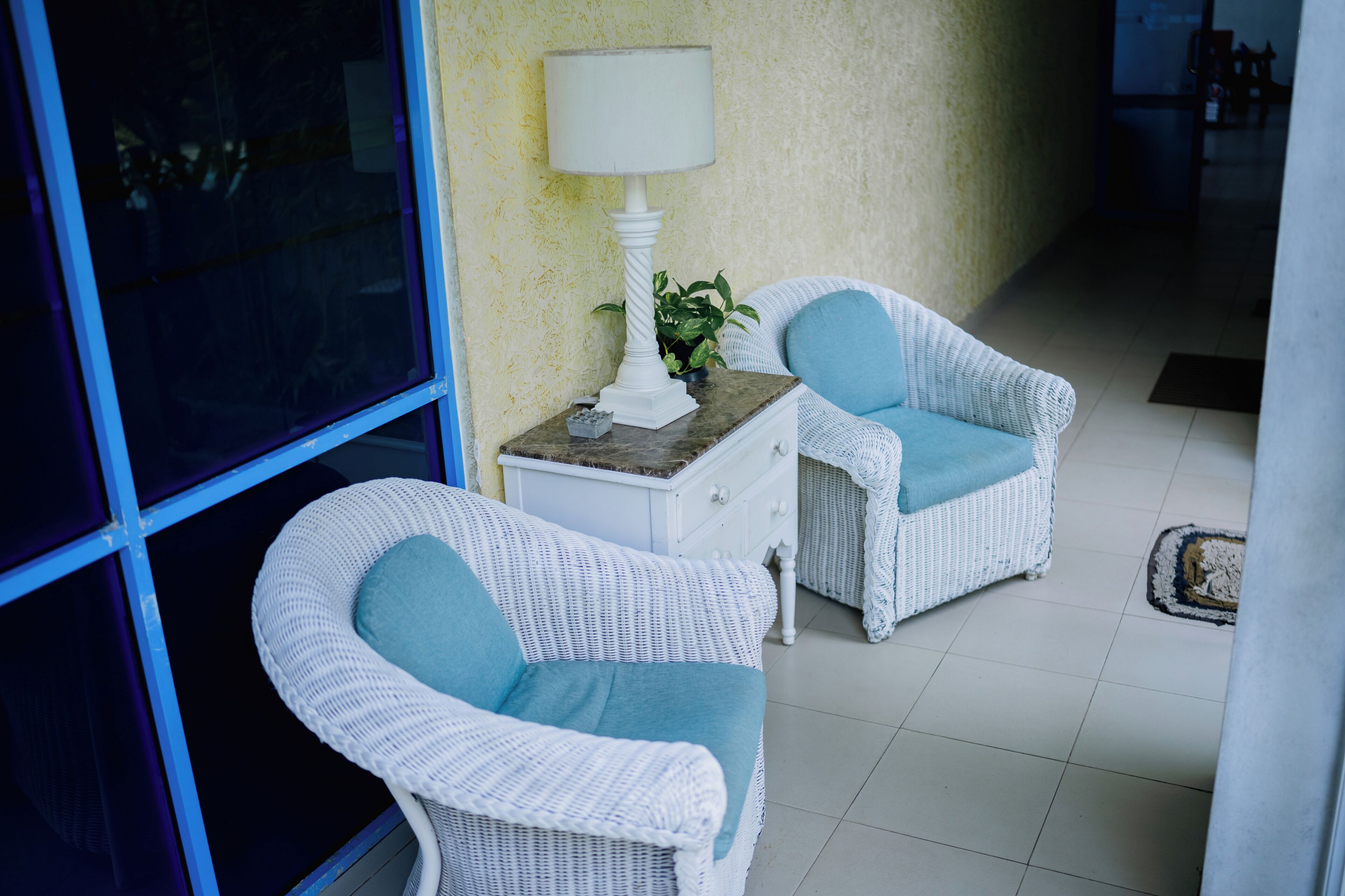 Two wicker chairs with blue cushions, a white lamp, and a small plant on a white table in a room with a yellow textured wall and a glass door.