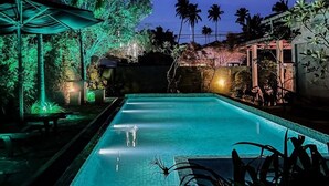 En pool som används för kvällsbad, omgiven av palmer, med en byggnad synlig i bakgrunden.