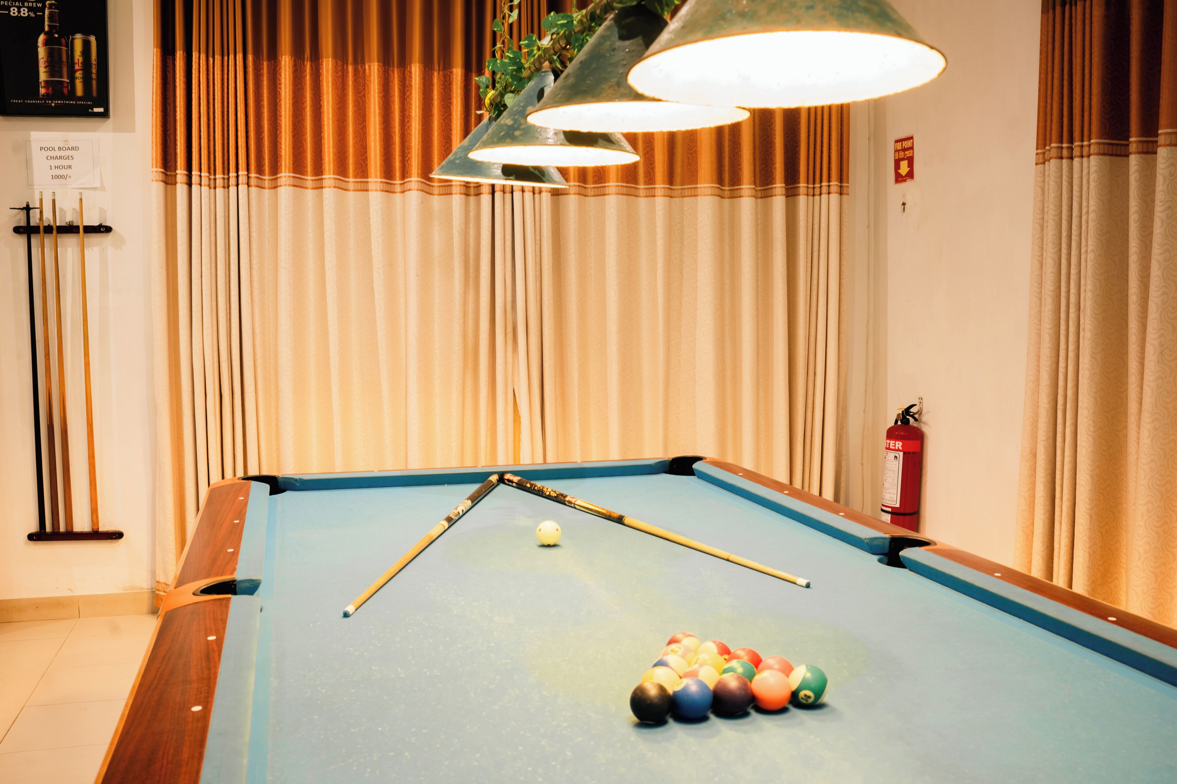 Een pooltafel met een keu, biljartballen en een brandblusser in een kamer met gordijnen en een bord dat aangeeft wat het gebruik van de pooltafel kost.