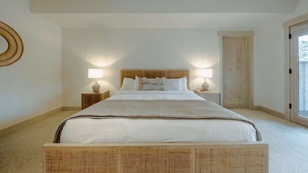 Un dormitorio con una cama grande, dos mesitas de noche con lámparas, un cabecero de madera y una puerta a la derecha.