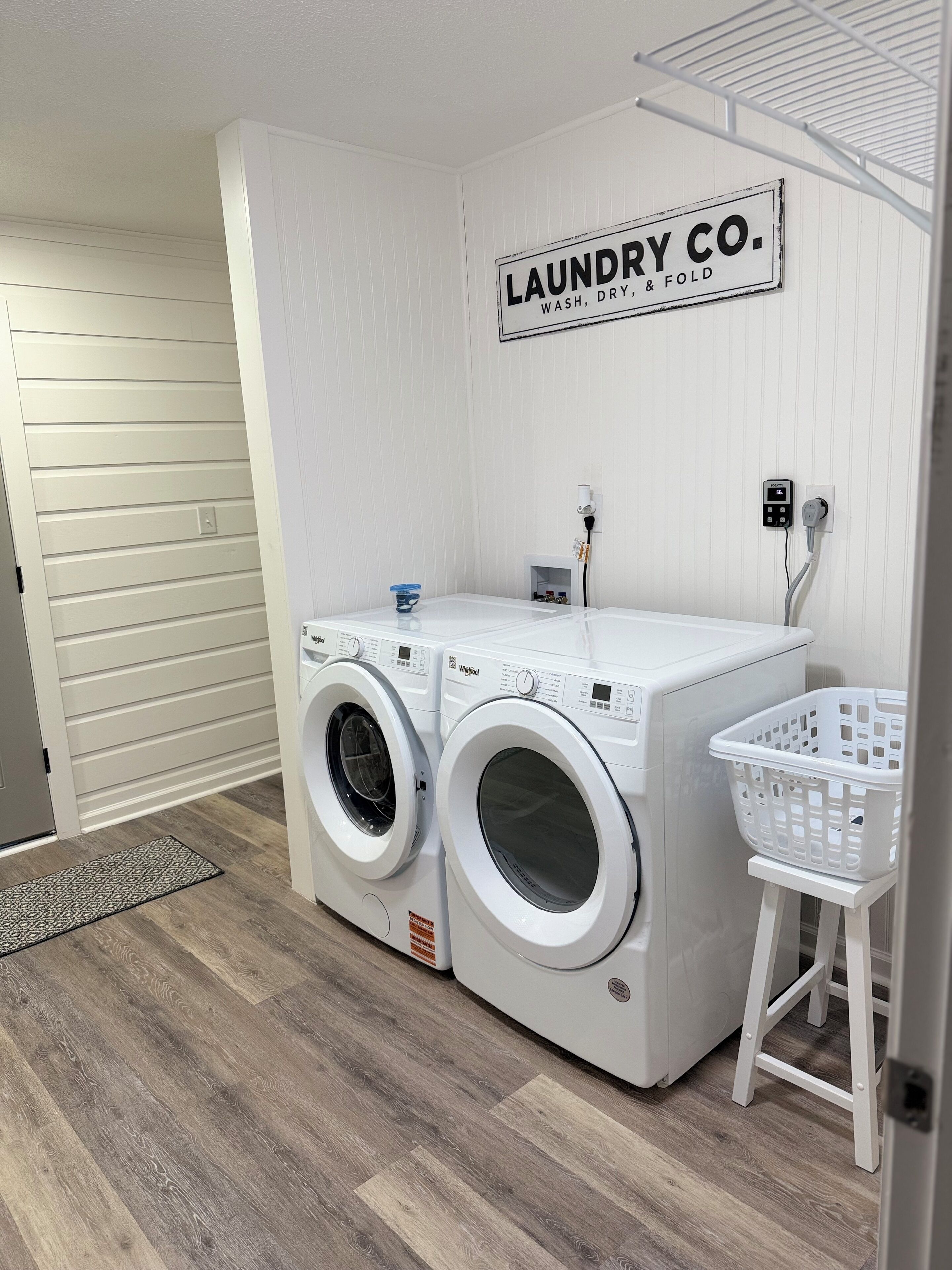 Un cuarto de lavandería con dos lavadoras de carga frontal, un cartel en la pared que dice "LAUNDRY CO. WASH, DRY, & FOLD", y una cesta de ropa blanca sobre un taburete.