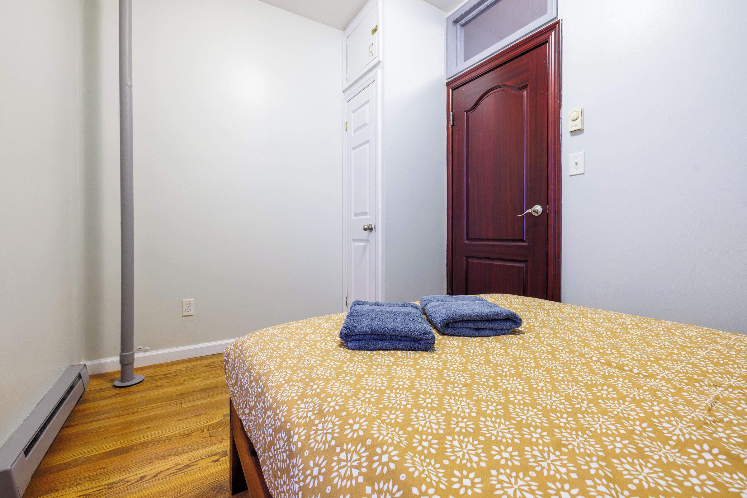 A bedroom with a bed, two towels, a wooden floor, a white wall, a white door, and a red door.