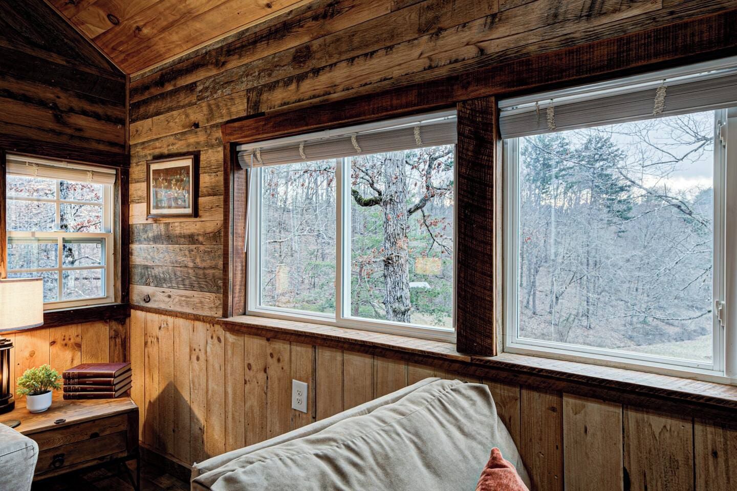 A cabin interior with wooden walls, a bed, a desk with books, and a window with a view of trees.