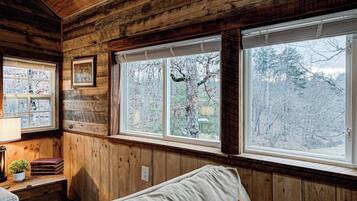 A cabin interior with wooden walls, a bed, a desk with books, and a window with a view of trees.
