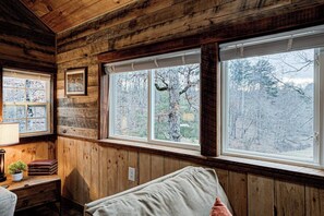 Interior de una cabaña con paredes de madera, una cama, un escritorio con libros y una ventana con vistas a árboles.
