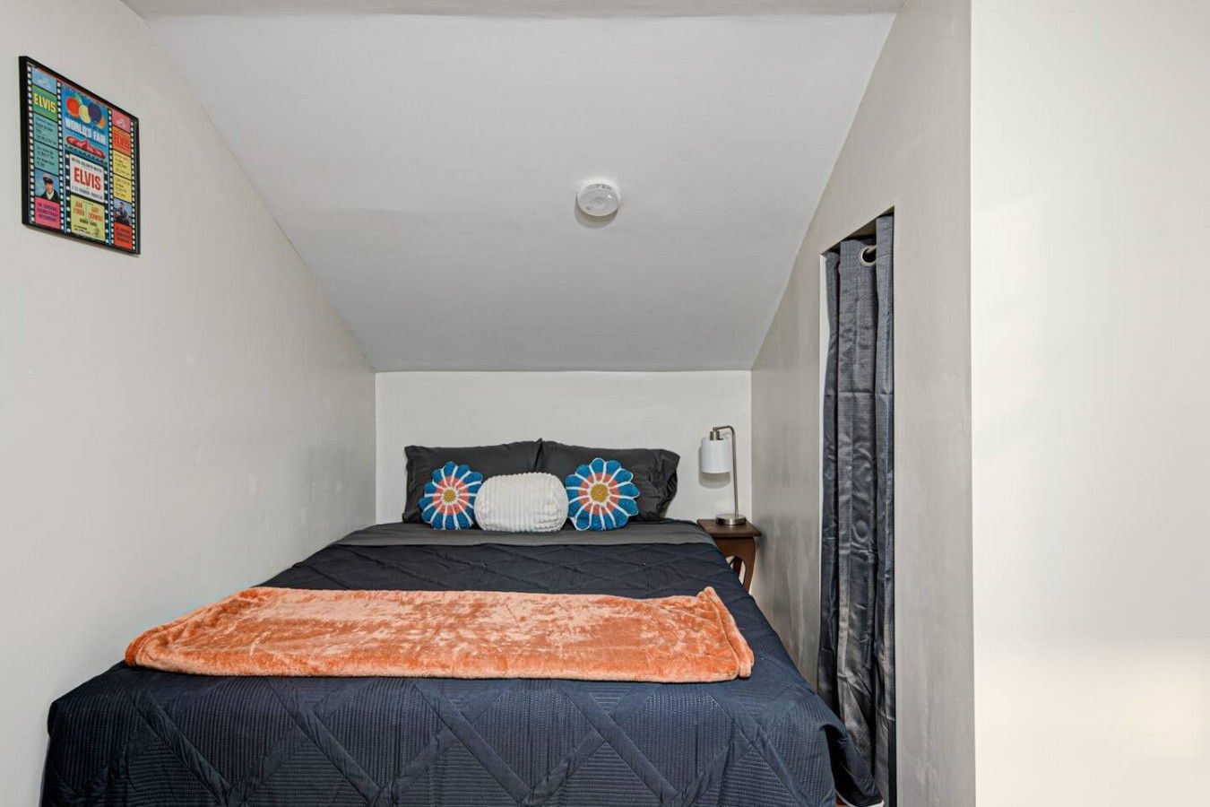 A bedroom with a bed, pillows, a blanket, a nightstand, a lamp, and a wall decoration.