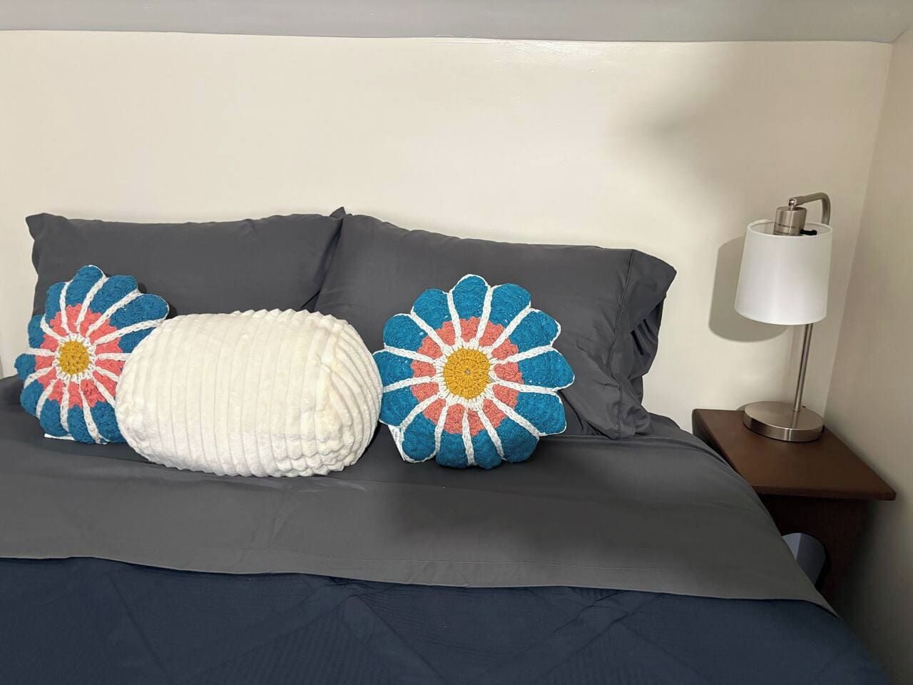 A bed with a grey duvet, two pillows with floral patterns, and a bedside lamp on a wooden nightstand.