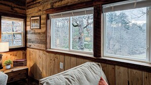 A cabin with wooden walls, a bed, a desk with a lamp, and a window with a view of trees.