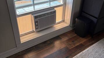 Una habitació amb una finestra gran, aire condicionat integrat, una cadira de color fosc i un terra de fusta.