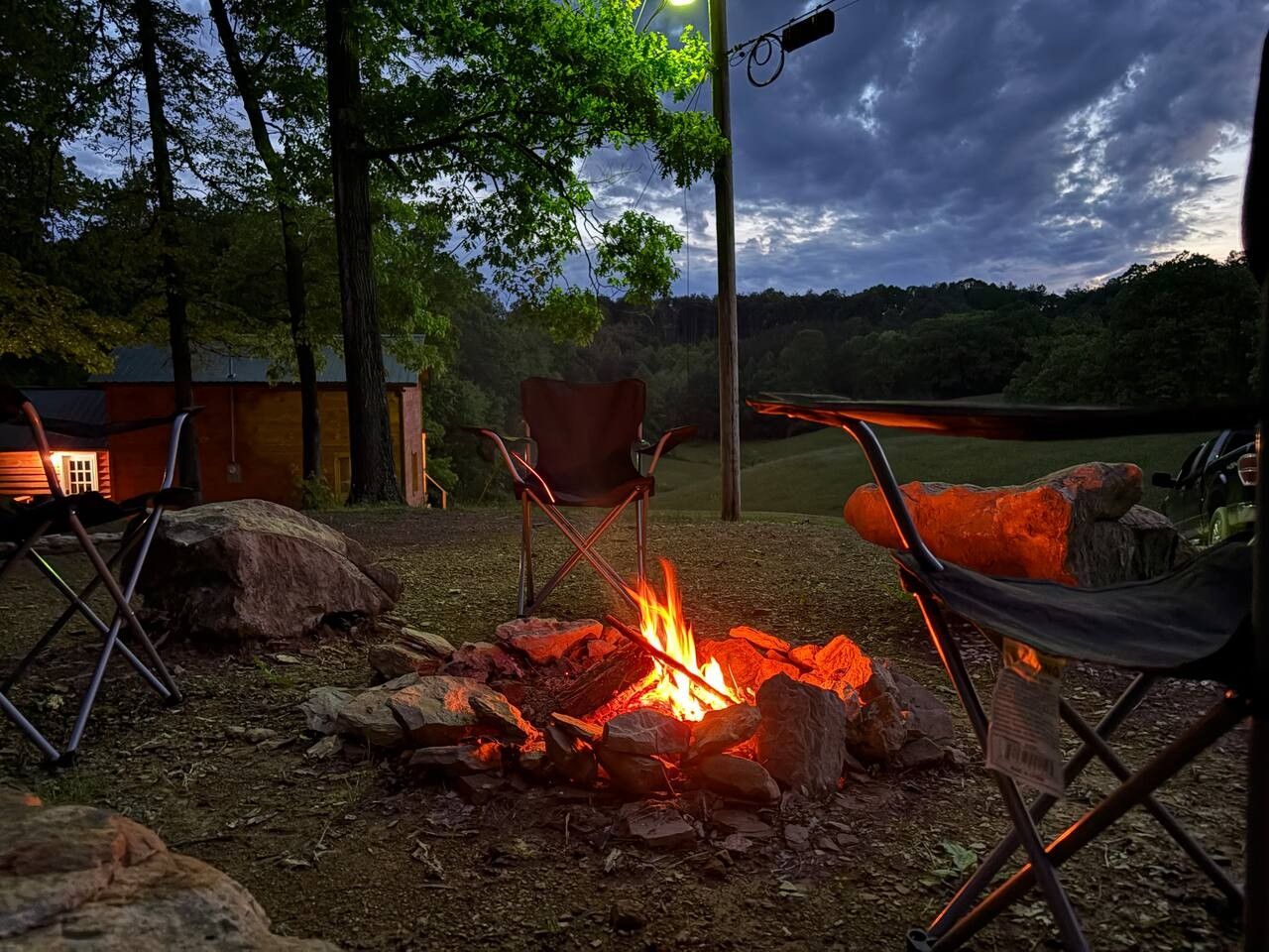 An outdoor seating area with a campfire, two folding chairs, and a rustic wooden structure in the background.