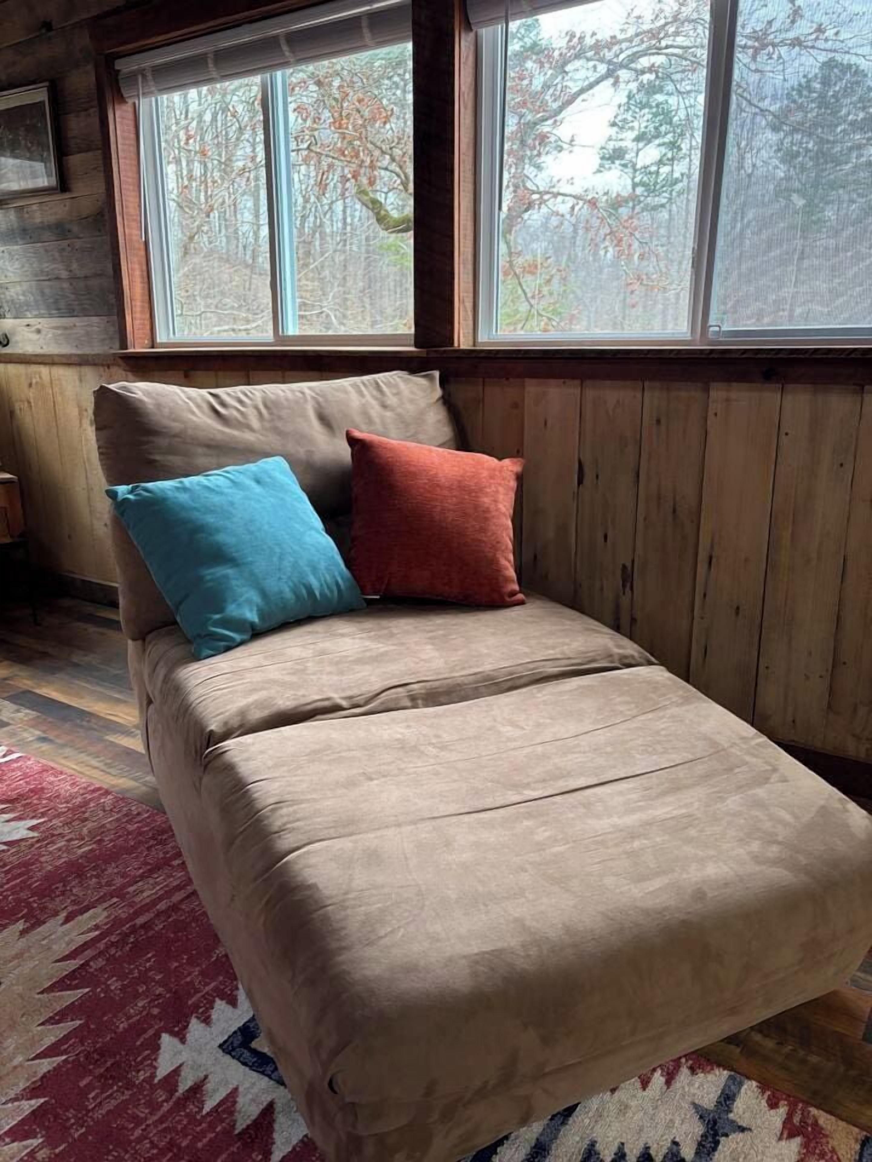 Un lit simple recouvert d’une couverture beige, deux oreillers, une fenêtre donnant sur des arbres et un mur en bois.