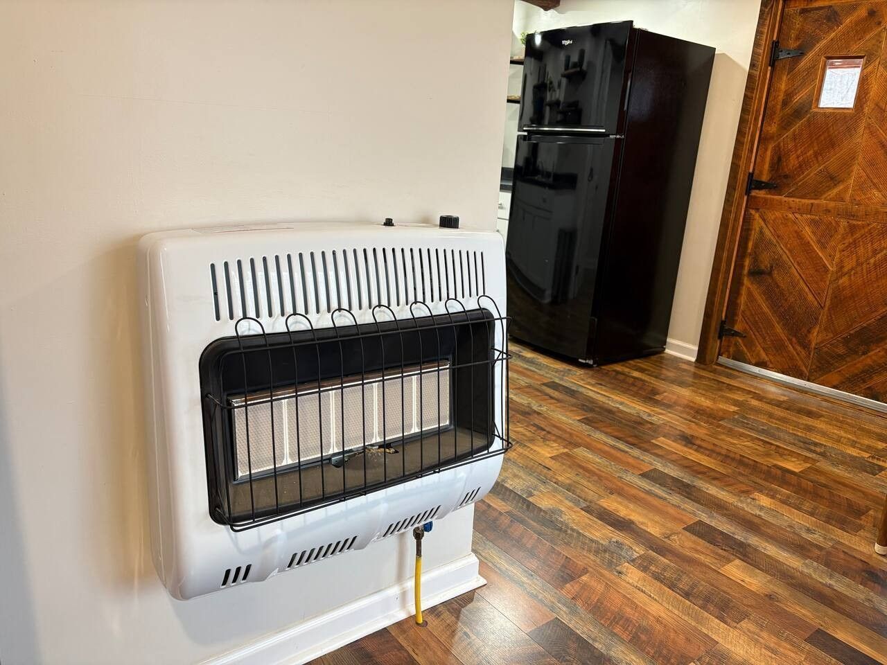 Un calentador blanco montado en la pared con rejilla de ventilación, un refrigerador negro y una puerta de madera en una habitación con suelo de madera.