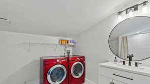 Une buanderie équipée de deux machines à laver à chargement frontal de couleur rouge, d’un évier blanc avec un miroir et d’une étagère au-dessus des machines.